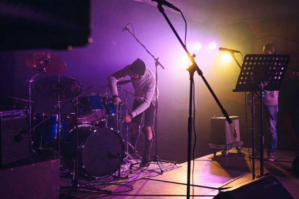 A small concert stage during setup with purple and yellow stage lighting, where a musician wearing a beanie adjusts a drum kit at center stage while microphones on stands, a music stand with sheet holes, an amplifier, and cables are positioned around the stage, creating a hazy live performance atmosphere before the show begins.